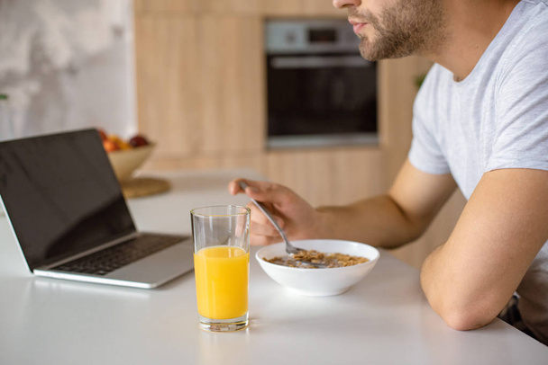cropped image of man eating flakes at kitchen table with fresh juice and laptop  - Photo, Image