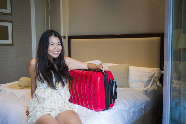 lifestyle portrait of young happy and beautiful Asian Korean tourist woman with travel suitcase just arrived at five star hotel room sitting on bed staring at balcony excited enjoying holidays trip  - Foto, imagen