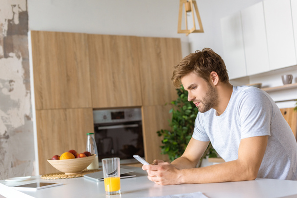serious young man using smartphone at kitchen table with juice and laptop  - Foto, Bild