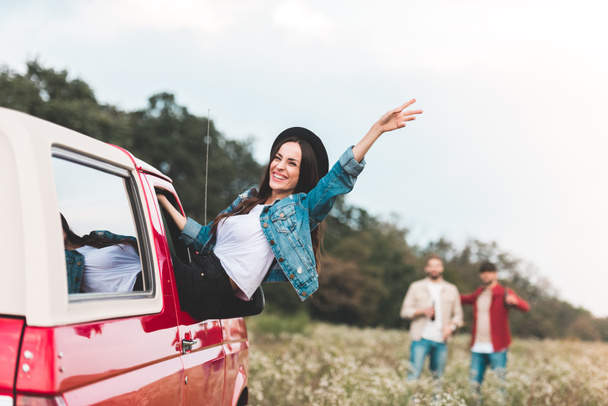 young woman outstretching from car window and raising hand while men standing blurred on background in flower field - Fotoğraf, Görsel