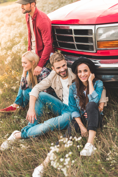 group of happy young car travellers sitting on flower field and leaning back on vintage truck - Photo, image