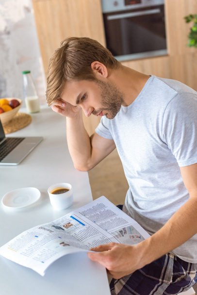 сосредоточенный человек, читающий газету за кухонным столом с кофе и ноутбуком
  - Фото, изображение