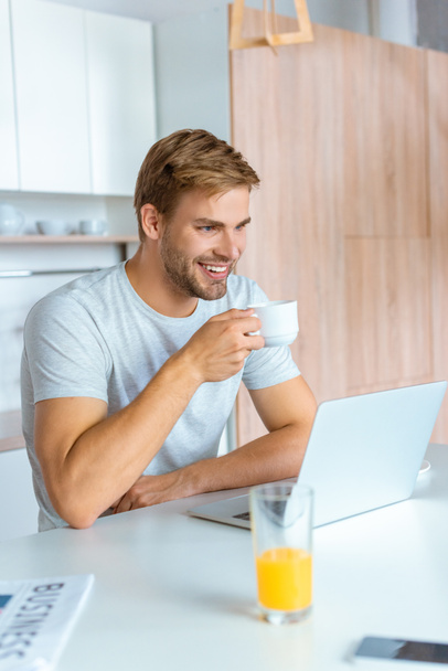 happy young man drinking coffee and looking at laptop screen at kitchen  - Foto, Imagem