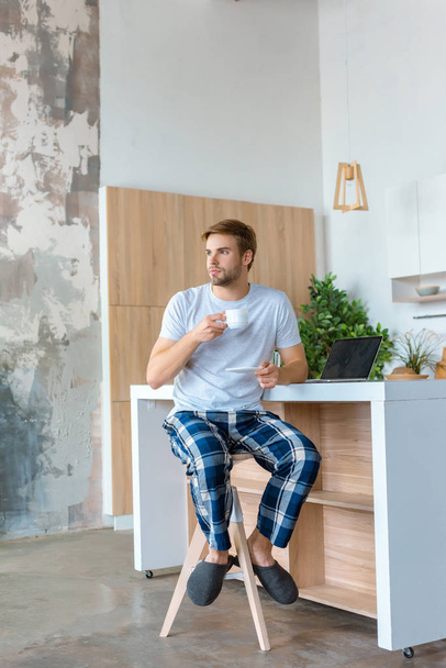 young man drinking coffee and looking away at table with laptop  - Foto, Imagen