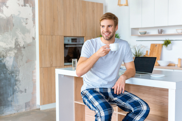 smiling young man drinking coffee at kitchen table with laptop  - Photo, image