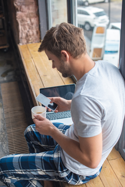 νεαρός άνδρας με την Κύπελλο Καφές χρησιμοποιώντας smartphone, ενώ κάθεται στο περβάζι στο σπίτι - Φωτογραφία, εικόνα