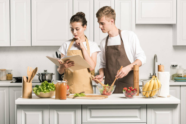 young couple cooking salad and reading recipe in cookbook in kitchen - Foto, imagen