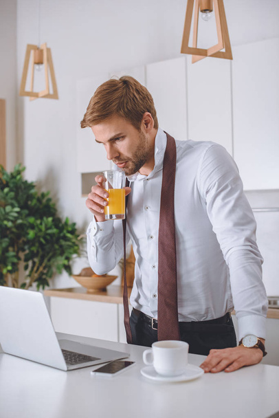 young businessman drinking fresh juice and looking at laptop screen at kitchen during morning time  - Foto, afbeelding