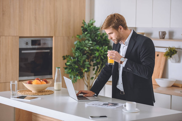 focused businessman drinking fresh juice using laptop at kitchen table during breakfast  - Fotó, kép