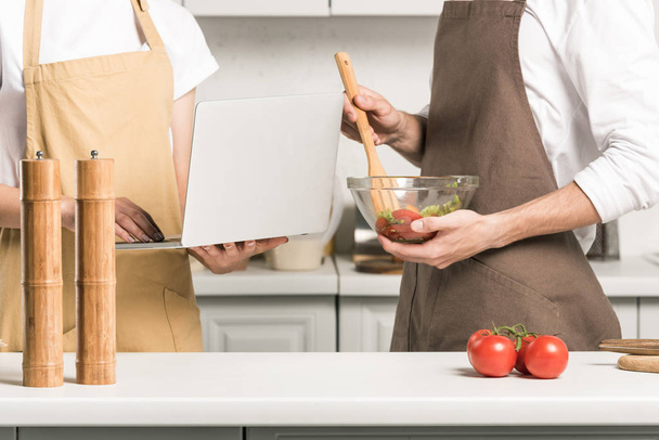 cropped image of couple cooking salad and using laptop in kitchen - Photo, image
