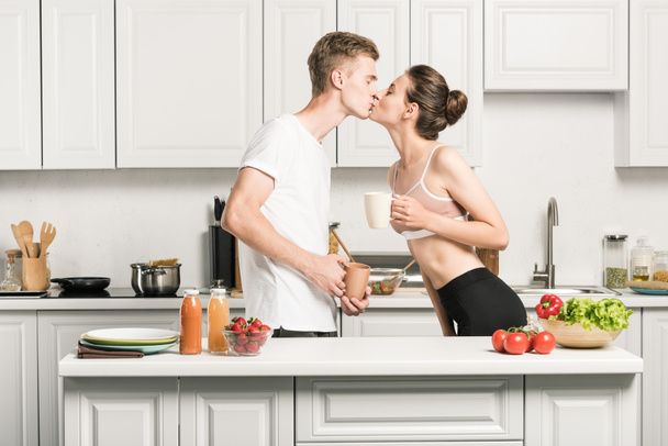 vista laterale della giovane coppia in possesso di tazze e baci in cucina
 - Foto, immagini