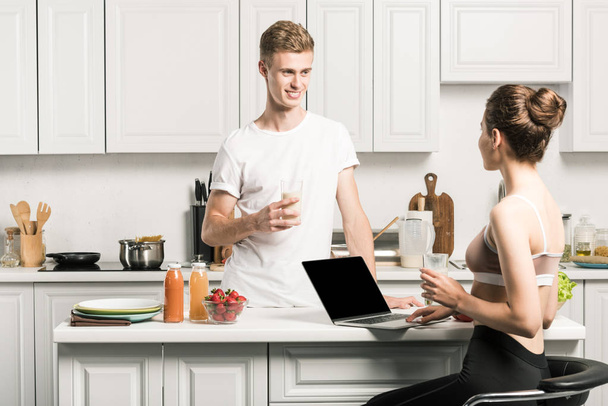 boyfriend holding glass of milk and girlfriend using laptop in kitchen - Photo, Image