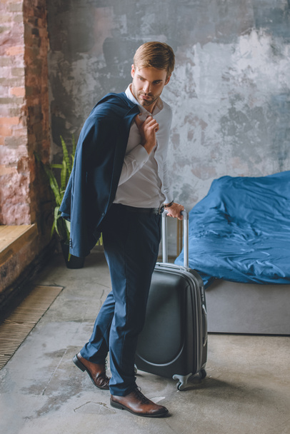 joven hombre de negocios con chaqueta sobre el hombro llevando maleta en el dormitorio en casa
 - Foto, Imagen