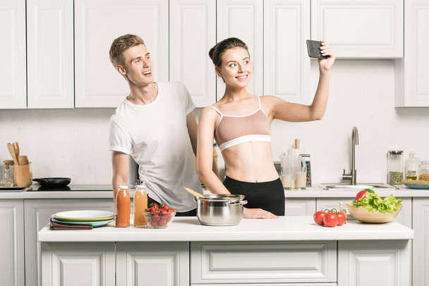 φιλενάδα σε αθλητικά σουτιέν λαμβάνοντας αυτοπορτρέτα με smartphone στην κουζίνα - Φωτογραφία, εικόνα