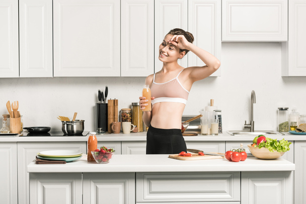 attractive girl in sport bra touching forehead and holding bottle of fresh juice in kitchen - Valokuva, kuva