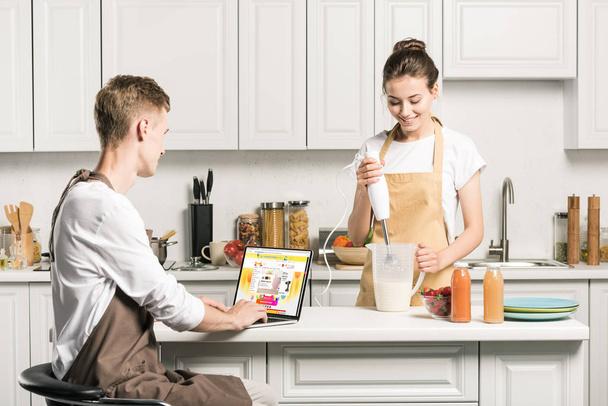 girlfriend cooking and boyfriend using laptop with loaded aliexpress page in kitchen - Foto, imagen