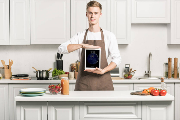 красавчик в фартуке, показывающий айпад с загруженной страницей на кухне
 - Фото, изображение