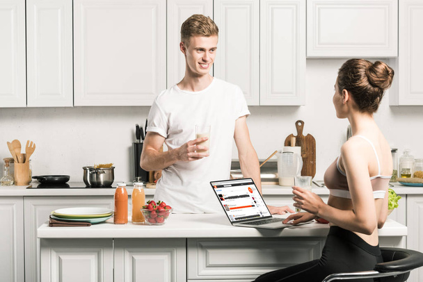 girlfriend using laptop with loaded soundcloud page and looking at boyfriend in kitchen - Фото, изображение