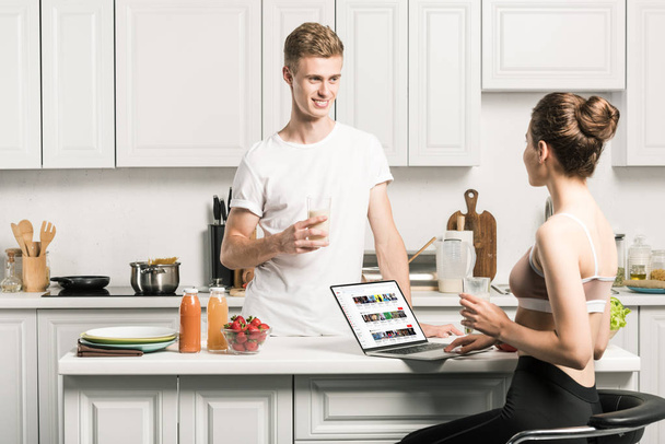 girlfriend using laptop with loaded youtube page and looking at boyfriend in kitchen - Foto, Imagem