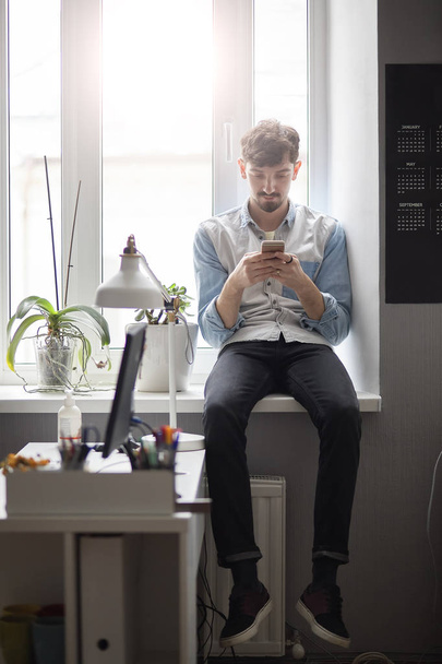 Elegantní mladík sedící na okně s telefonem v ruce - Fotografie, Obrázek