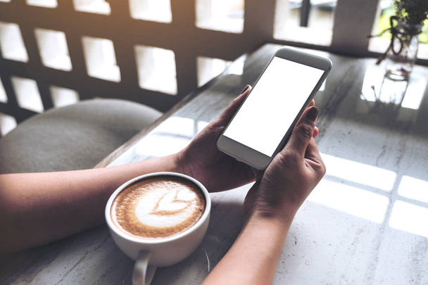 Imagem Mockup das mãos da mulher segurando telefone celular branco com tela de desktop em branco e xícara de café na mesa no café
 - Foto, Imagem