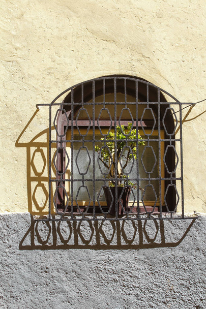 Bir kemer ve koyu renkli ahşap çerçeve penceresiyle. Kafes ve gölge. Tencere bitkiler. Sarı ve gri cephe. El Jadida, Morocco. - Fotoğraf, Görsel