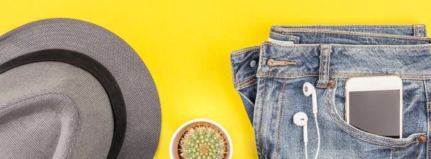 Плоская укладка синих джинсов, серая шляпа, кактус и наушники смартфона на жирном желтом бумажном фоне с копировальным пространством. Вид сверху на женщину в повседневной одежде. Длинное широкое знамя
 - Фото, изображение