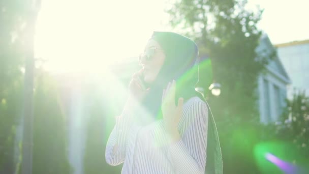 Young stylish attractive muslim woman in hijab and glasses talking on the phone,sunflare.Slow mo - Imágenes, Vídeo