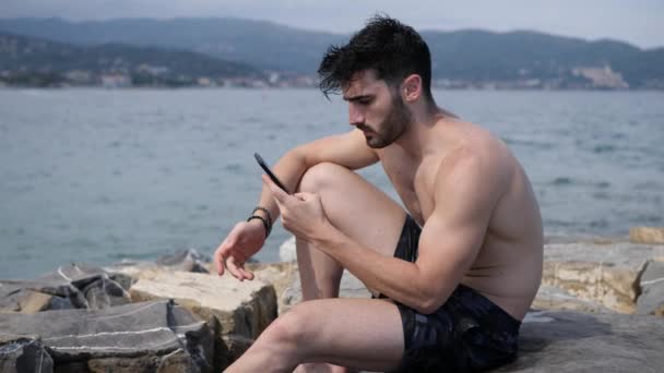Young man by the sea sending voice message on phone - Imágenes, Vídeo