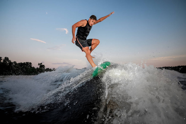 Homem ativo e jovem pulando no wakesurf para baixo das ondas do rio contra o céu azul ao pôr do sol
 - Foto, Imagem