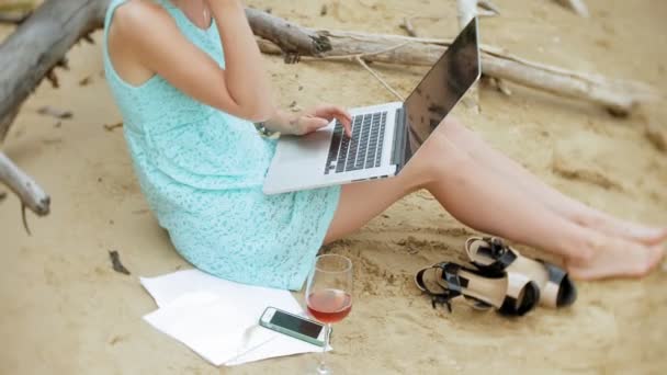 drukke mooie jonge vrouw die werken op een laptopcomputer met handelspapieren oppakken van een telefoon om te werken een oproep zittend op het strand aan zee op een zonnige dag en drinken wijn uit een glas - Video