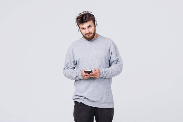 A young man with a beard in everyday clothes listens to pleasant music in wireless headphones. Gray background with space for text - Φωτογραφία, εικόνα