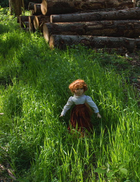 Beautiful vintage doll with red hair standing in sunlight, in green grass, in the forest alone - Photo, Image