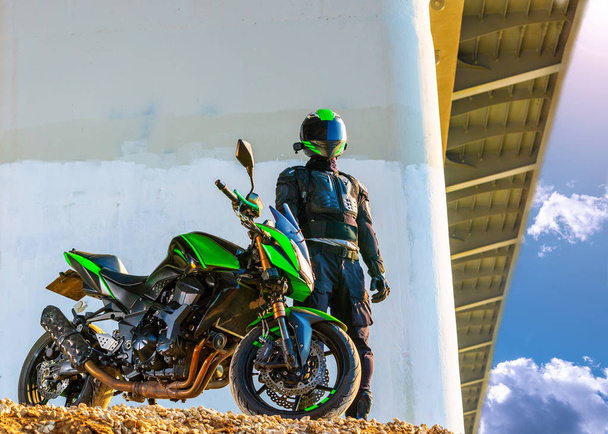 moottoripyöräilijä kypärässä ja suojapuvussa seisoo sillan alla
 - Valokuva, kuva