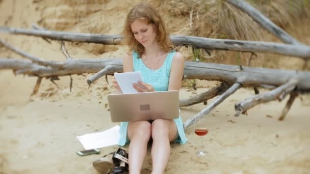busy beautiful young woman working on a laptop computer with business papers picking up a phone to work a call sitting on the beach by the sea on a sunny day and drinking wine from a glass - Footage, Video