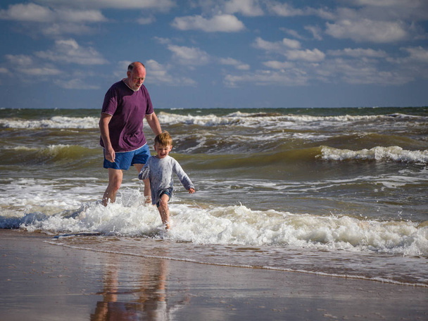 Vitaler bärtiger Opa und verspielter Enkel mit nassen Klamotten läuft mit Freude am Strand im Meerwasser. - Foto, Bild
