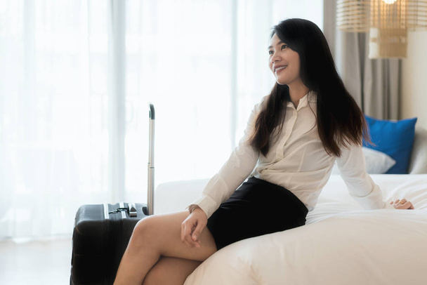 Smiling Asian business woman sitting on bed in hotel room. Business travel concept - Valokuva, kuva