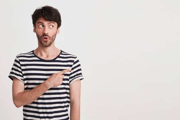 Portret van verbaasd knappe jongeman met borstelharen draagt gestreepte t shirt voelt zich afgevraagd en wijst naar de kant met vinger geïsoleerd op witte achtergrond - Foto, afbeelding