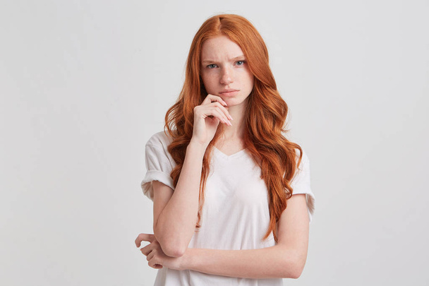 Portrait de jeune femme inquiète coûteuse aux longs cheveux roux ondulés et aux taches de rousseur garde les mains pliées et pense à ses problèmes isolés sur fond blanc
 - Photo, image
