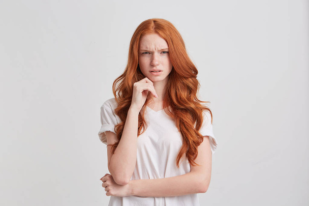 Closeup of upset unhappy redhead young woman with long wavy hair and freckles wears stylish t shirt keeps hands folded and looks angry isolated over white background - Foto, afbeelding