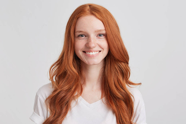 Closeup χαρούμενα όμορφη κοκκινομάλλα κοπέλα με μακριά κυματιστά μαλλιά και φακίδες φοράει πουκάμισο t φαίνεται αυτοπεποίθηση και αισθάνεται ευτυχισμένος απομονωθεί σε λευκό φόντο κοιτάζει απευθείας στην κάμερα - Φωτογραφία, εικόνα
