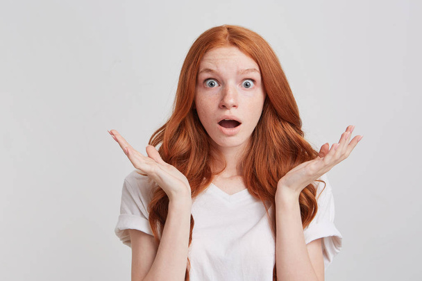 Портрет удивленной шокированной молодой женщины с длинными рыжими волнистыми волосами и открытым ртом носит футболку чувствует себя пораженным и выглядит удивленным изолированным на белом фоне
 - Фото, изображение