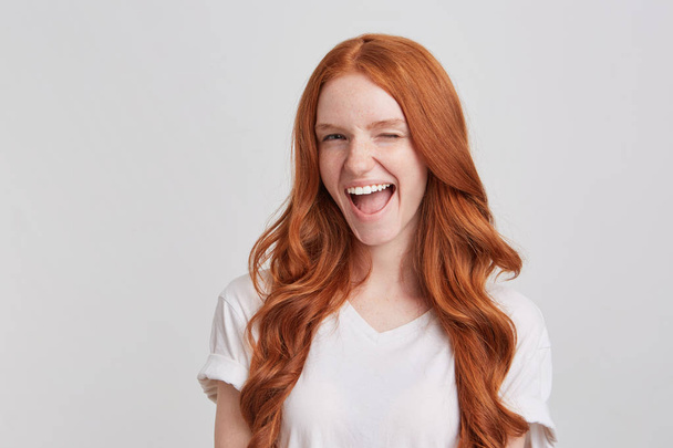 Close-up van cheeful speelse roodharige jonge vrouw met lang golvend haar draagt t shirt voelt gelukkig en succesvol en tevreden winking geïsoleerd op witte achtergrond kijkt - Foto, afbeelding