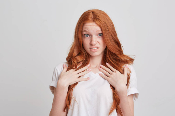 Porträt einer erstaunt unzufriedenen rothaarigen jungen Frau mit langen Haaren und Sommersprossen, die T-Shirt trägt, sieht erstaunt aus und zeigt auf sich selbst isoliert vor weißem Hintergrund - Foto, Bild
