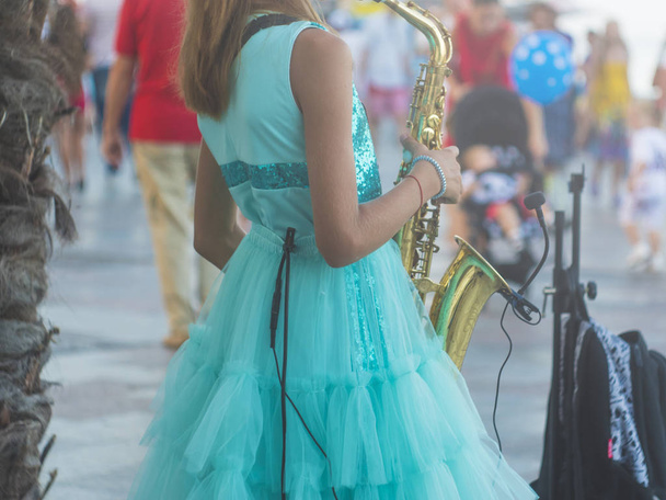 На заднем плане маленькая девочка, стоящая на многолюдной улице и играющая саксофонную музыку
 - Фото, изображение
