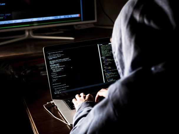 hacker in hoodie typing code in program using laptop in dark studio - Foto, afbeelding