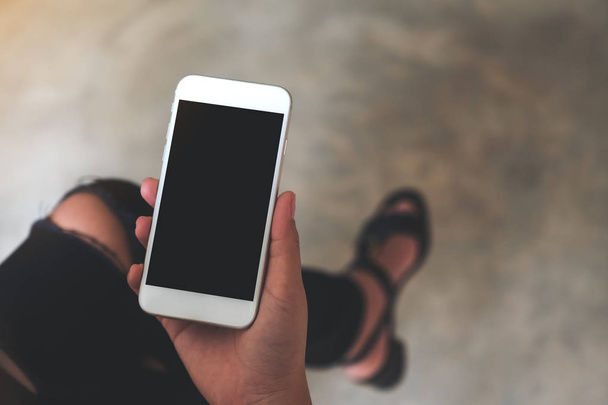 Top view mockup kép egy nő kezét tartó fehér mobiltelefon üres fekete képernyő a combon  - Fotó, kép