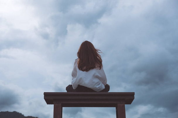 Nainen istuu yksin puisella penkillä puistossa pilvinen ja synkkä taivas tausta - Valokuva, kuva