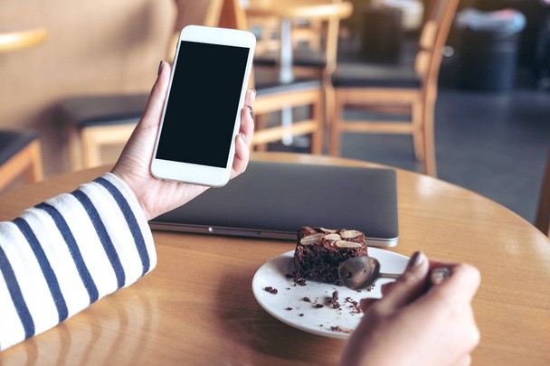 Mockup image of a woman holding white mobile phone with blank black desktop screen while enjoy eating brownie cake in cafe - Photo, Image