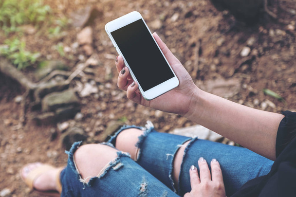 Imagen burlona de una mujer sosteniendo un teléfono móvil blanco con pantalla de escritorio en blanco mientras está sentada en el suelo al aire libre
 - Foto, Imagen